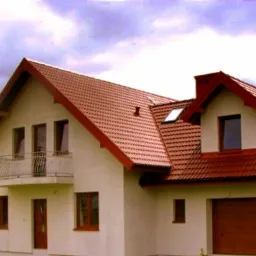 Dwupiętrowy dom z czerwonym dachem z dachówki, białymi ścianami i balkonem z metalową balustradą, garaż z brązową bramą.